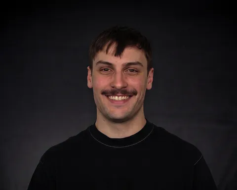 Junger Mann mit dunklem Haar und Schnurrbart lächelt vor schwarzem Hintergrund, trägt ein dunkles Shirt.