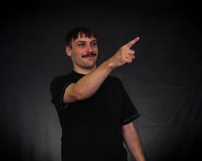 Mann zeigt mit dem Finger nach rechts und lächelt vor dunklem Hintergrund Mann mit Schnurrbart in schwarzem T-Shirt zeigt mit dem rechten Arm nach oben rechts vor schwarzem Hintergrund.