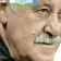 Älterer Mann mit grauem Schnurrbart und ernster Miene, Kopf ist seitlich zu sehen, unscharfer Hintergrund.